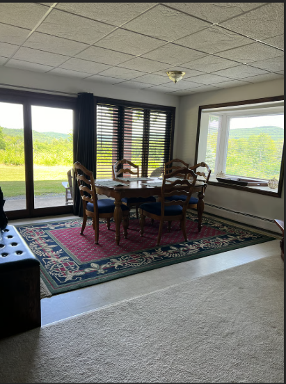 Dining area with wooden table and chairs in a room with large windows showing a view of greenery.