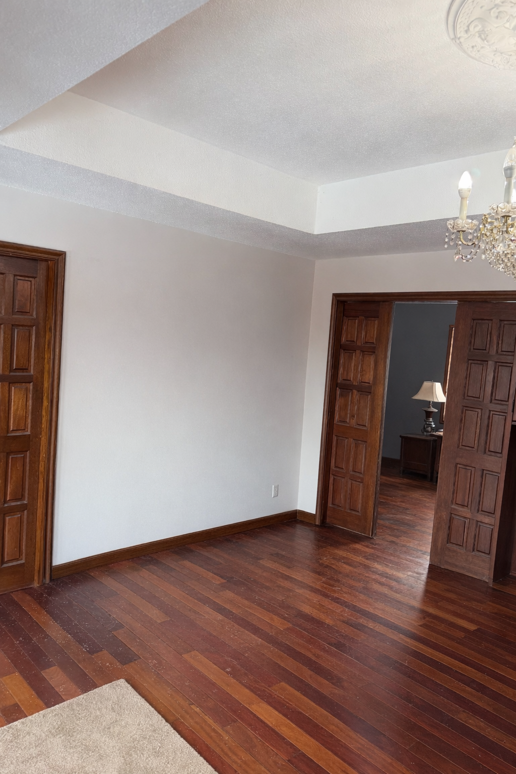Room with wooden  cherry floors and  double  sliding mahogany doors, chandelier, and lamp. 