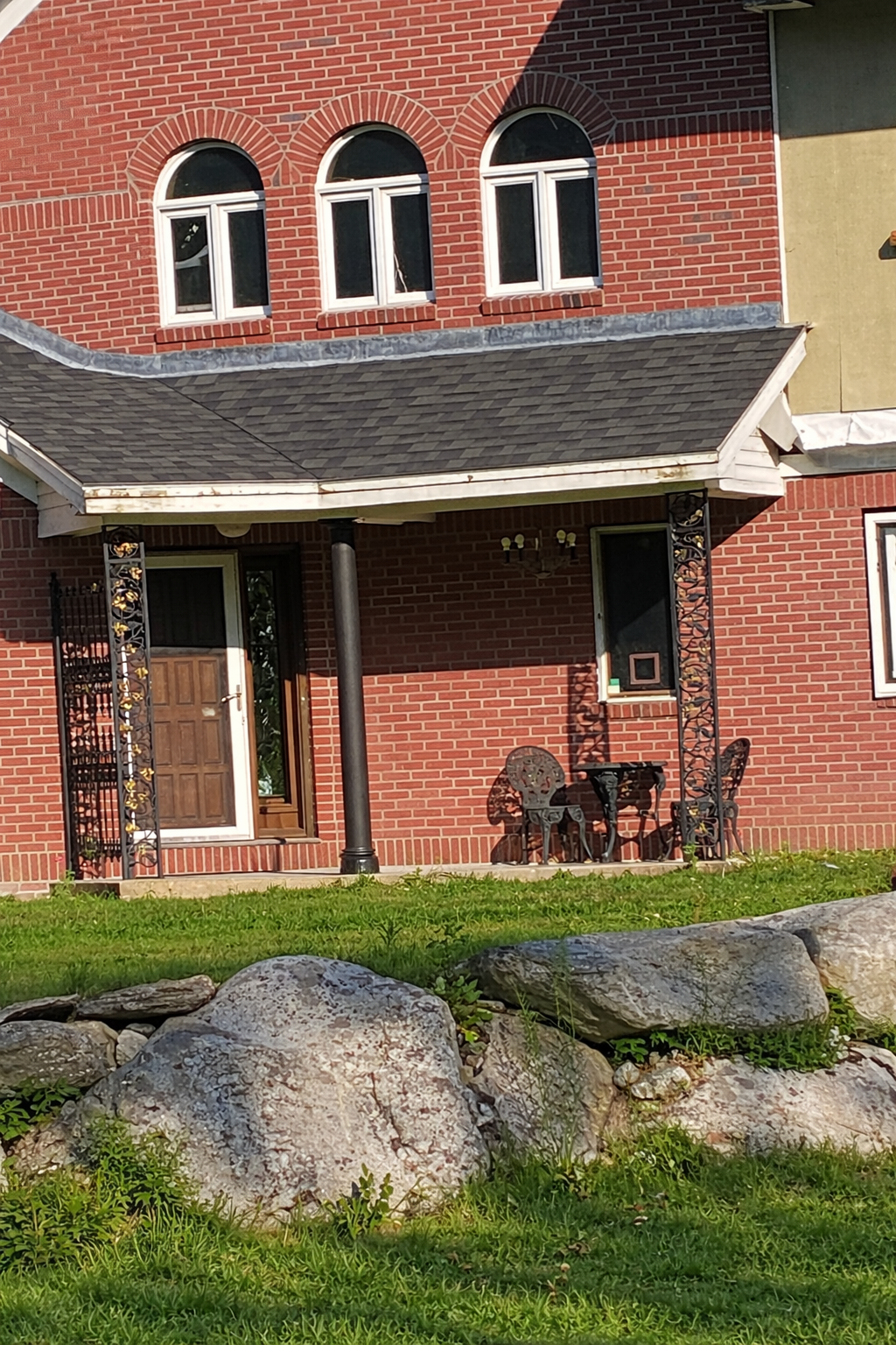 Red brick building with a porch and outdoor wrought iron  furniture on a grassy area.