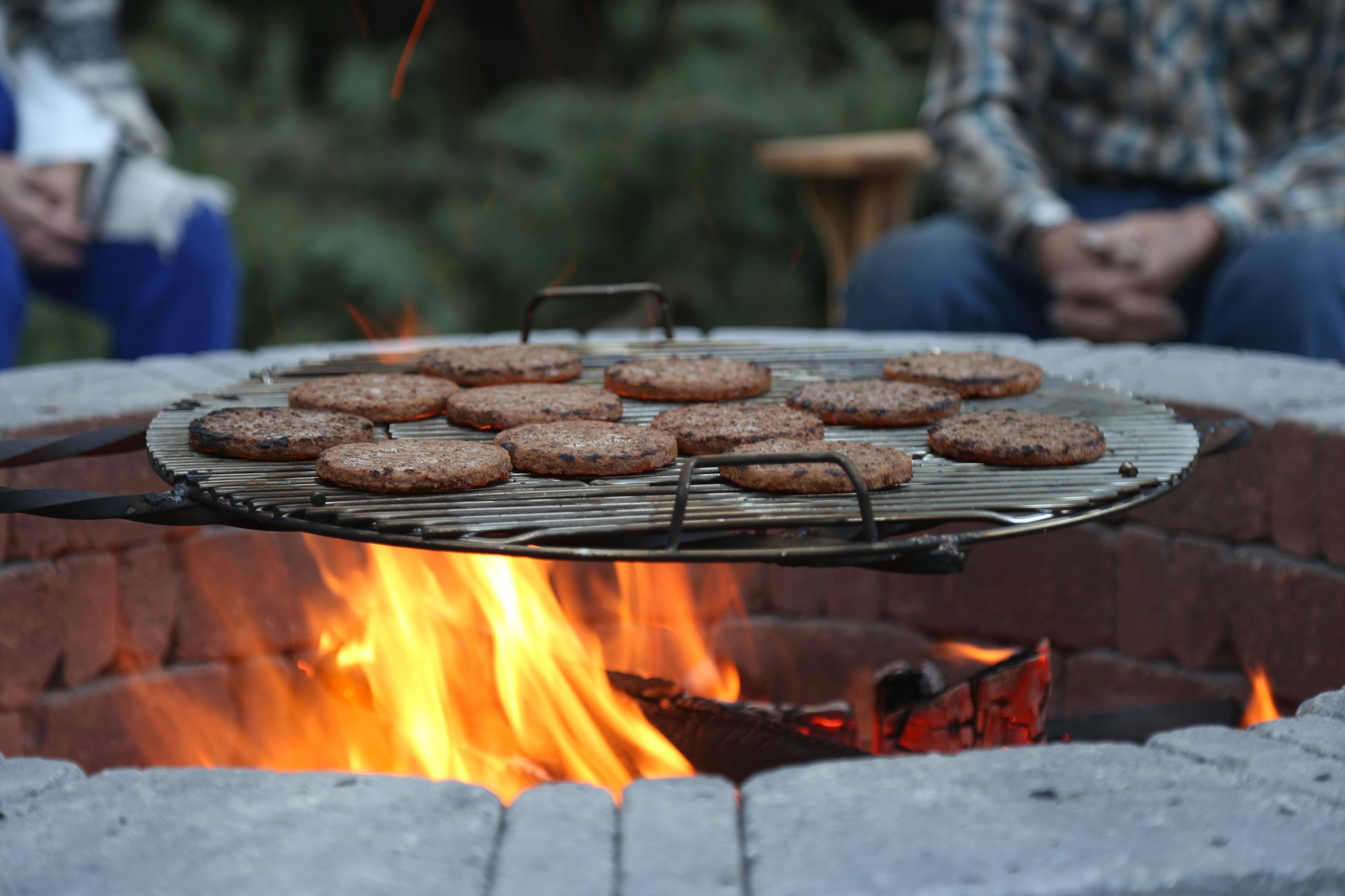 Hamburgers cooking on a grill over an open fire with people sitting around.