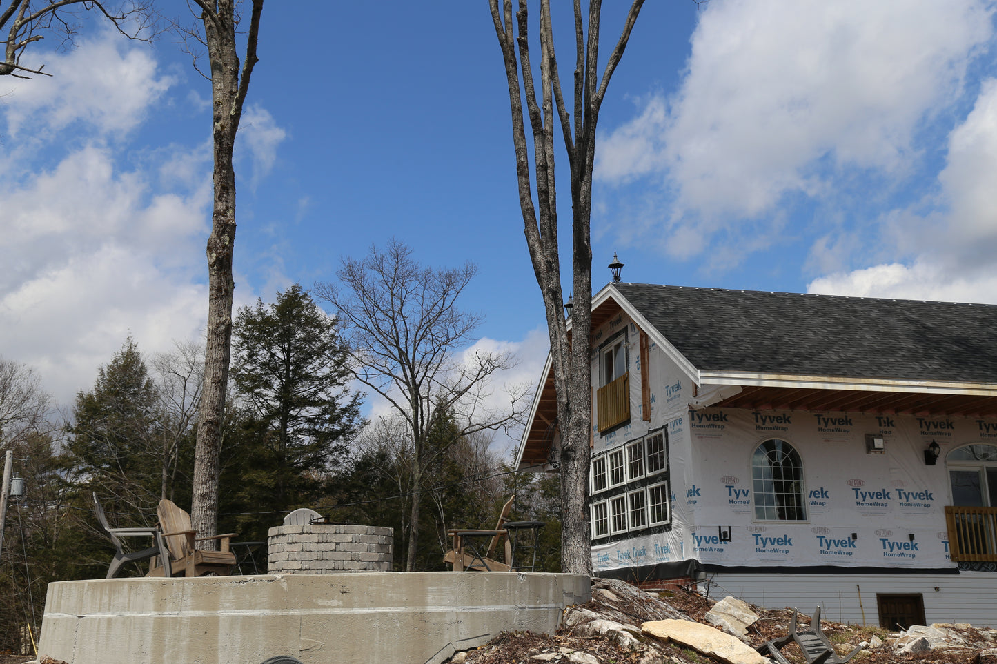 House under construction with Tyvek wrap on exterior walls, surrounded by trees and a clear sky.