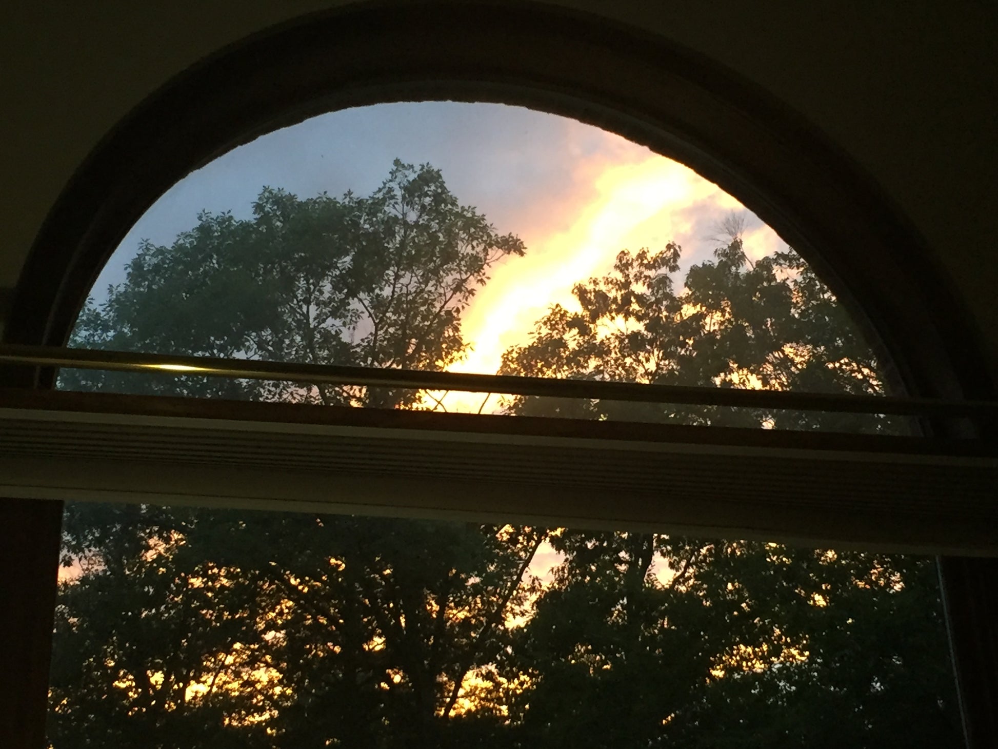 Window view of a rainbow arching over trees during sunset.