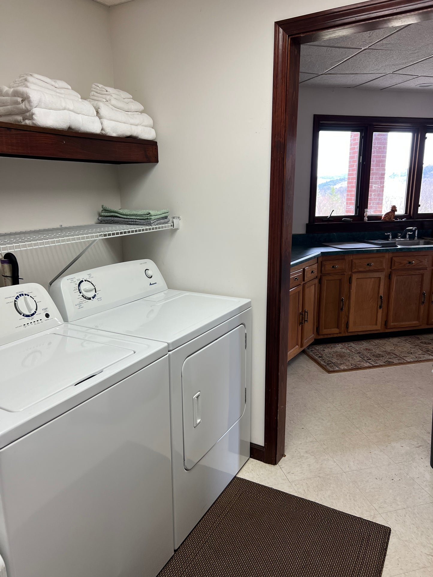 Laundry room with white washer and dryer, shelves with folded towels, and a view of a kitchen. Nature View NH Estate