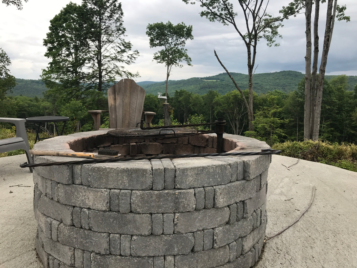 Stone fire pit with Adirondack chairs in a scenic outdoor setting with trees and mountains.