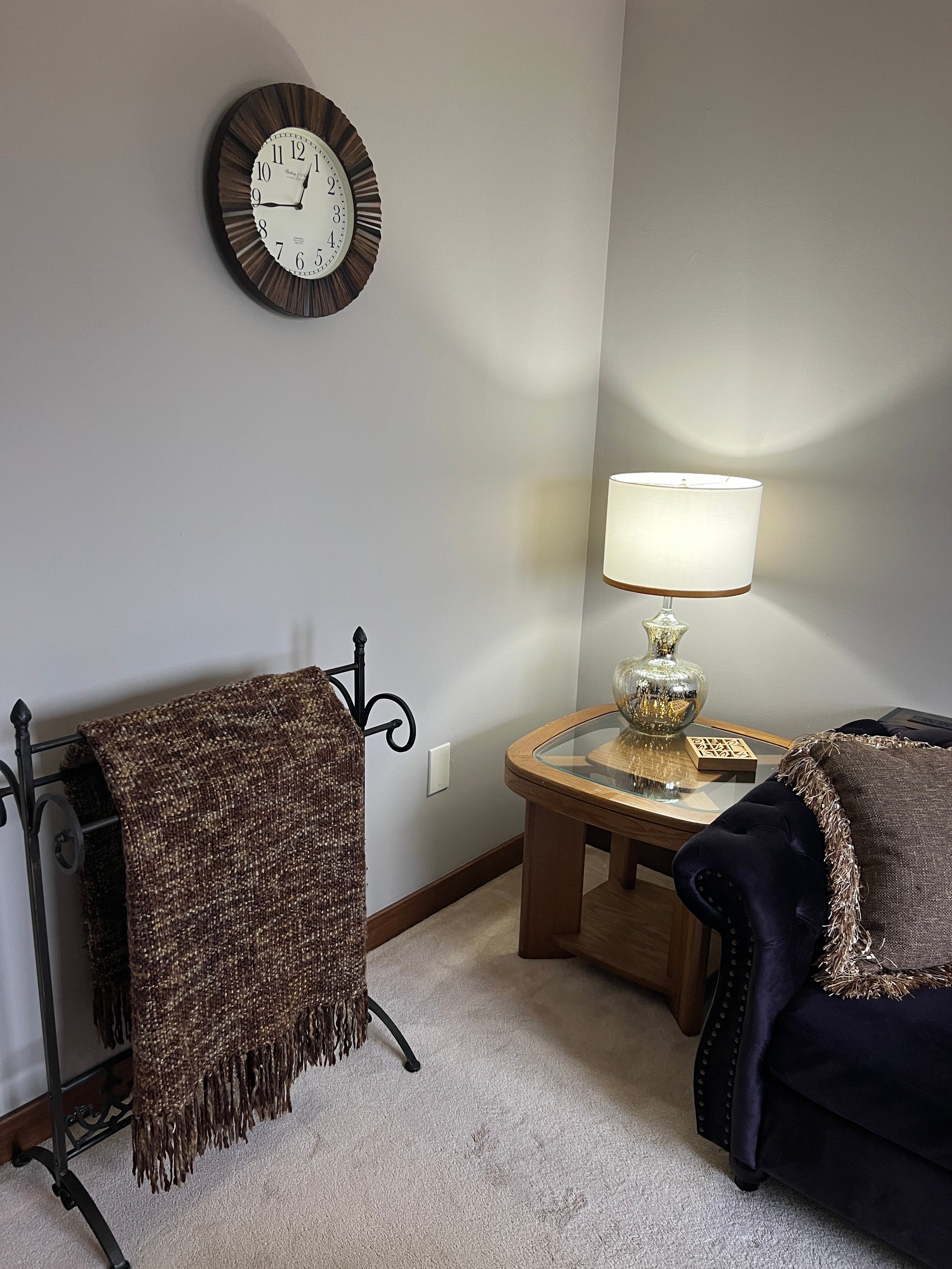 Small room with a clock on the wall, a lamp, and a chair with a blanket.