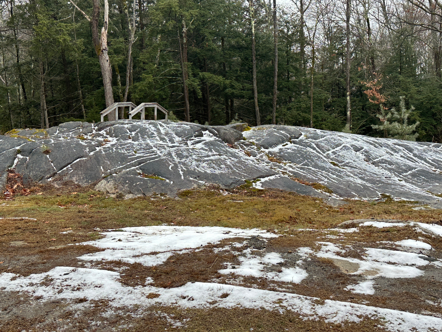 Rocks with patches of snow in a forested area bridge in background