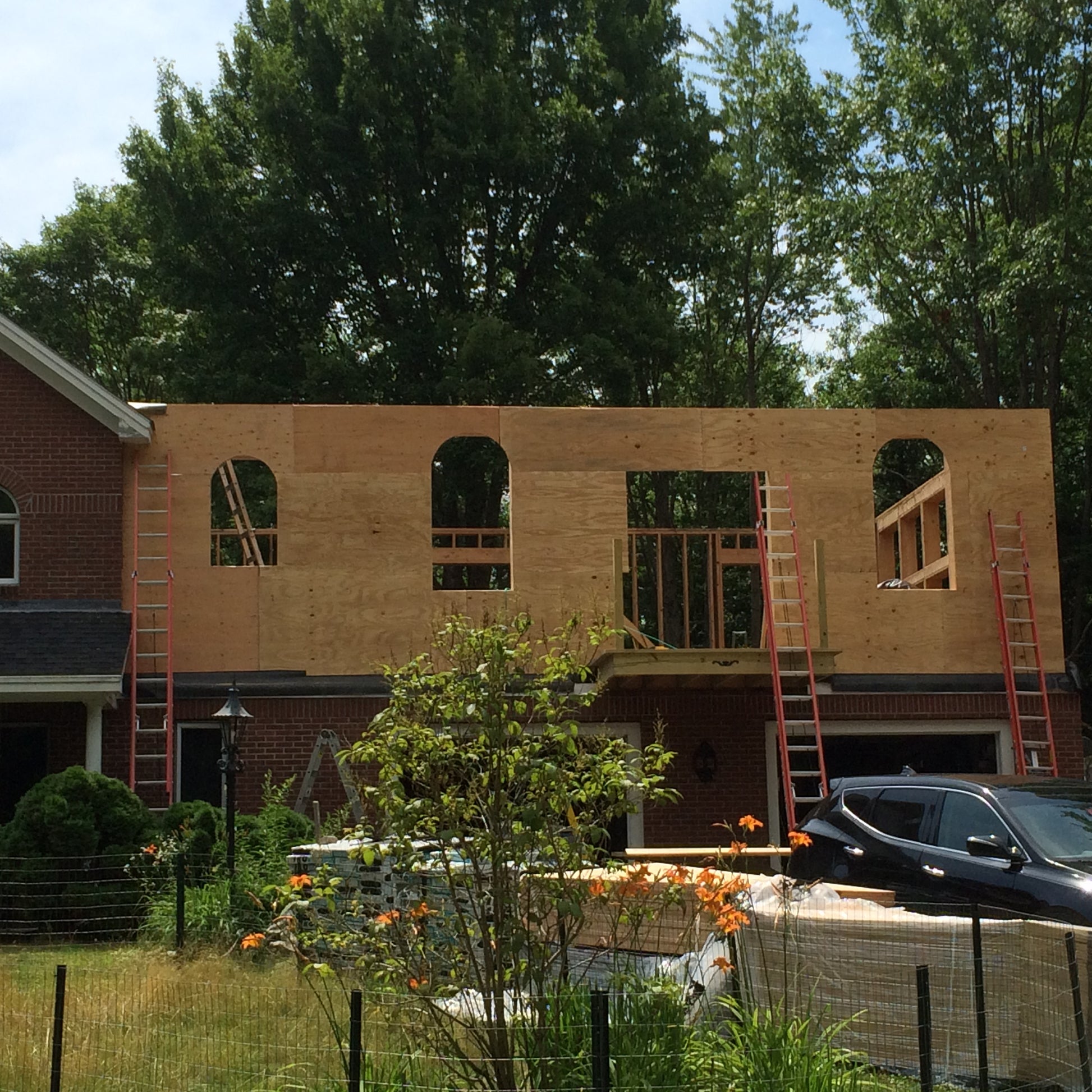 House under construction with wooden frame and visible windows, surrounded by trees and a car.