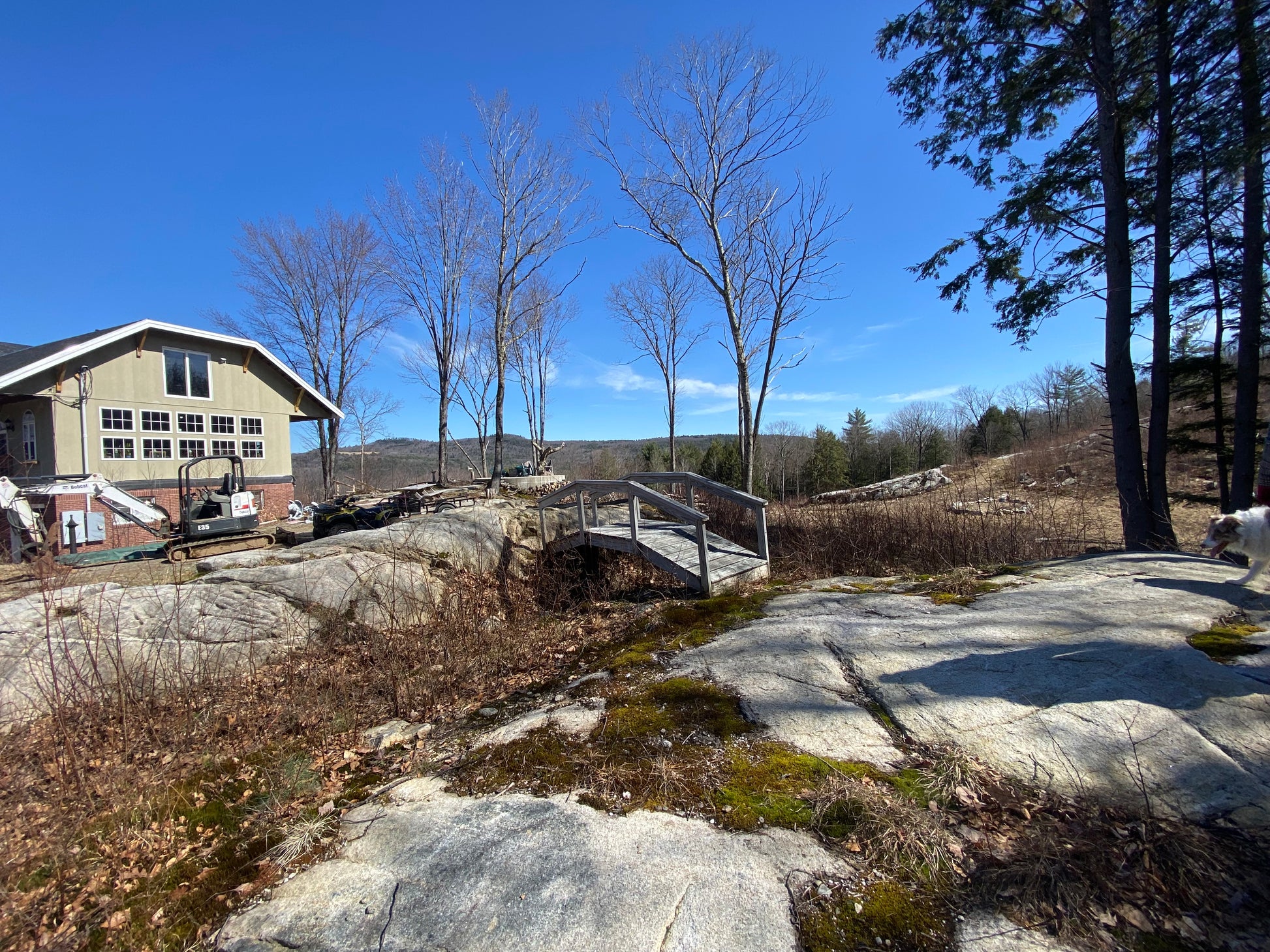 Rural landscape with a house, trees, and rocky terrain under a clear blue sky.