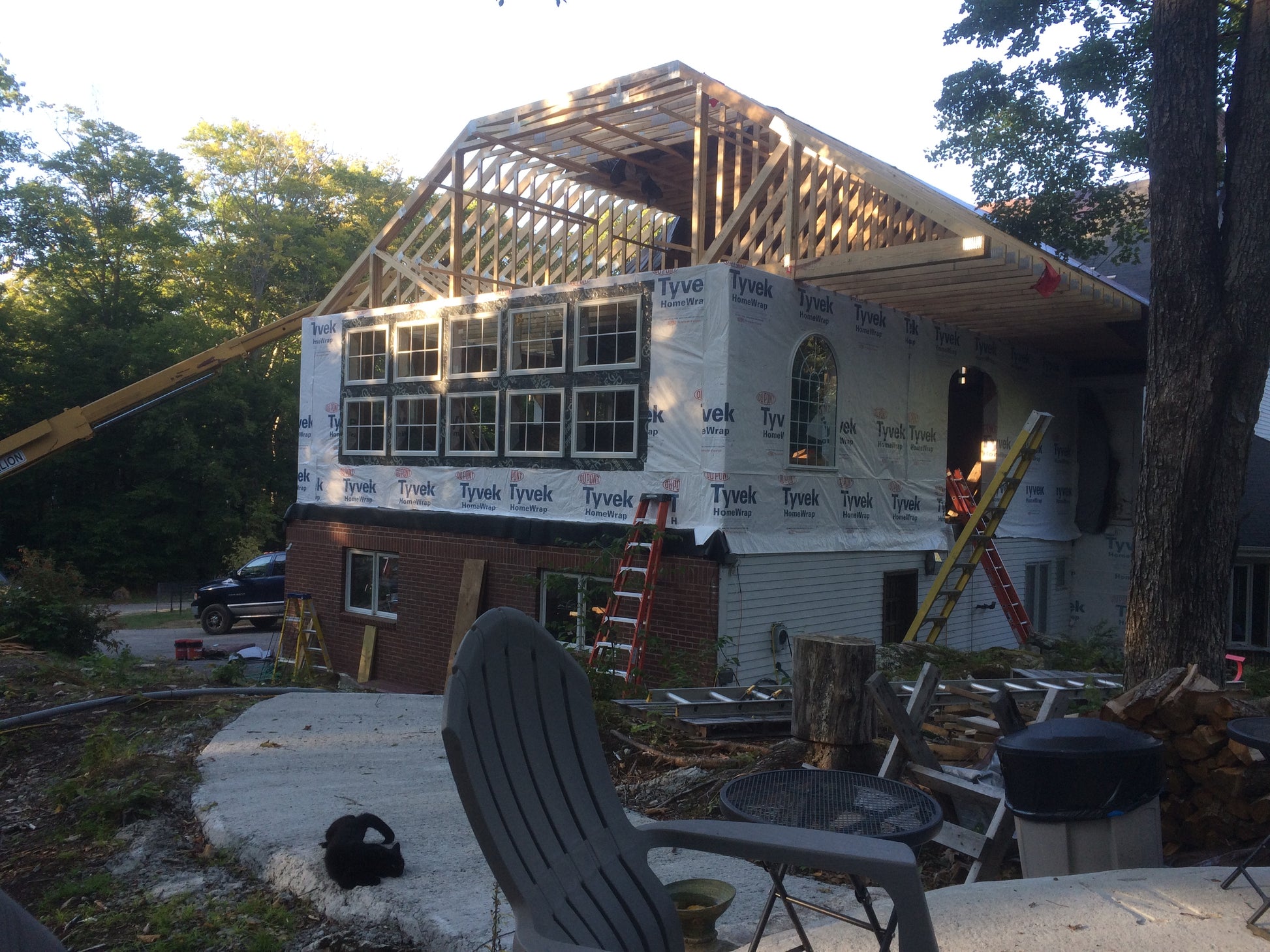 House under construction with wooden frame and Tyvek wrapping, surrounded by trees and outdoor furniture.