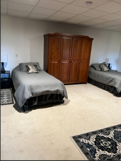 Two single beds with gray bedding and a wooden wardrobe in a room with beige carpet and white walls.