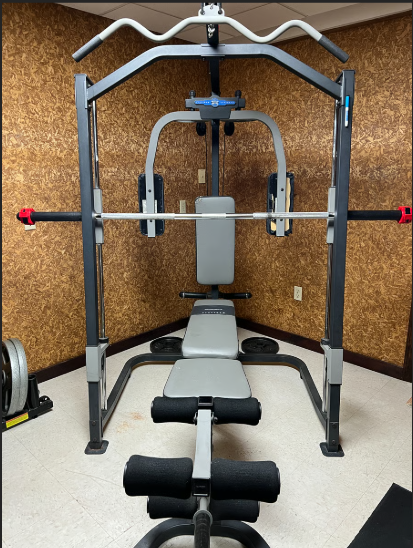Home gym equipment including a weight bench and barbell rack against a cork wall.